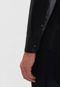 Braccio destro di una persona che indossa una camicia nera a maniche lunghe con polsino abbottonato, appoggiato naturalmente contro i pantaloni neri.