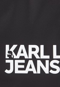 Melns audums ar baltām paceltām burtiem, kas rakstīti "KARL JEANS", ar nelielu reljefu vīrieša siluetu ar saulesbrillēm, kas integrēts burta "K".