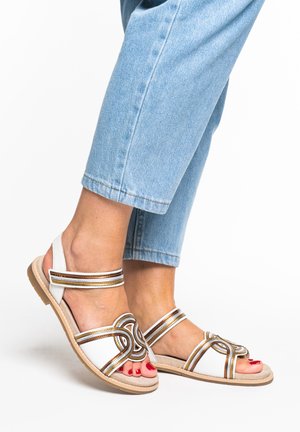 Feet wearing white sandals with metallic bronze straps and red nail polish, paired with light blue cropped jeans against a white background.