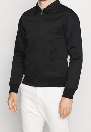 Homme portant une veste noire zippée avec une poche à rabat boutonnée et un pantalon blanc sur un fond clair uni.