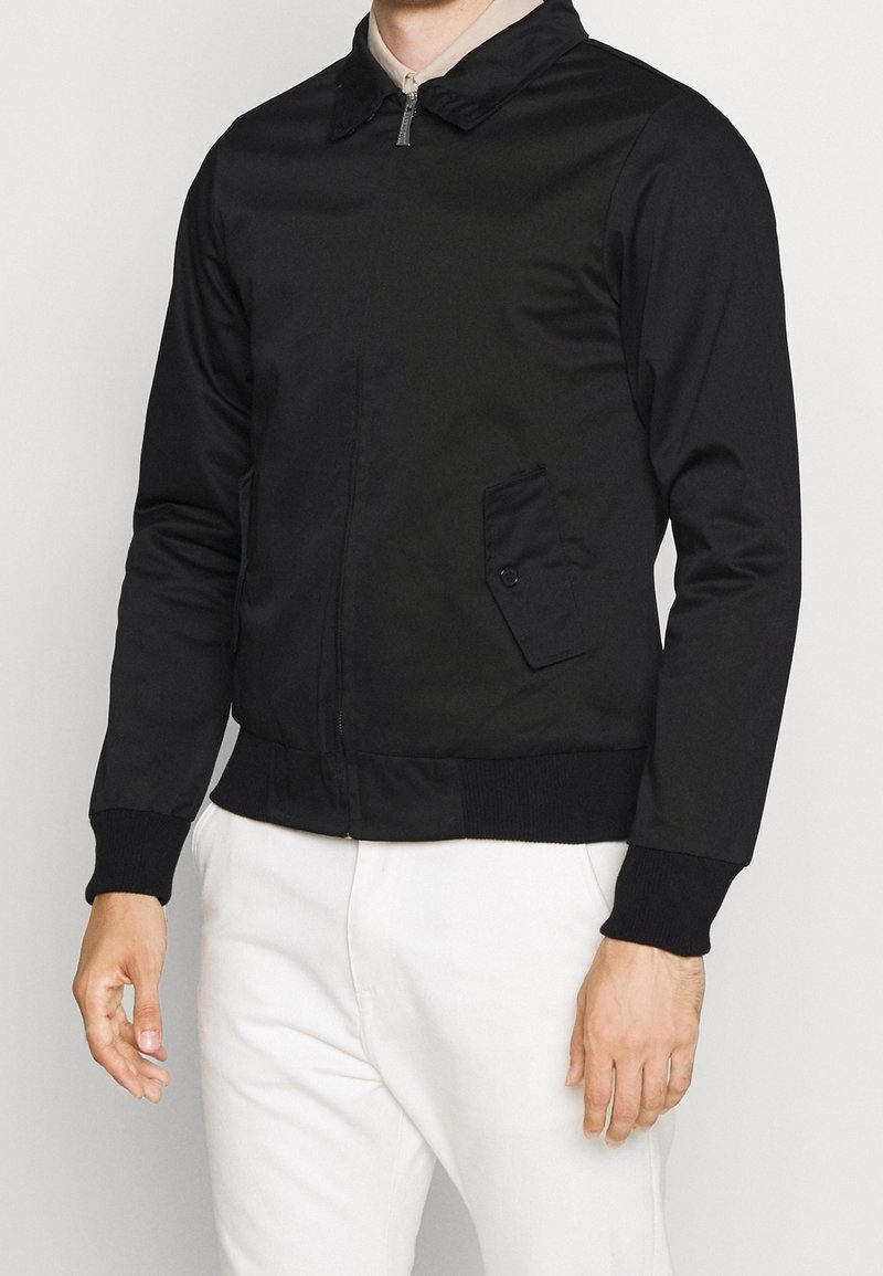 Homme portant une veste noire zippée avec une poche à rabat boutonnée et un pantalon blanc sur un fond clair uni.