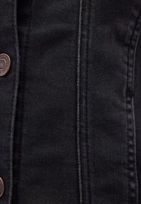 Tessuto in denim nero con cuciture visibili e bottoni in tonalità rame. La texture appare liscia con un design classico.