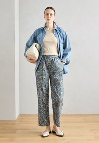 Giacca di jeans sopra un top beige, pantaloni blu a fantasia floreale e ballerine color crema. Tiene una borsa color crema e sta in piedi su un pavimento di legno.