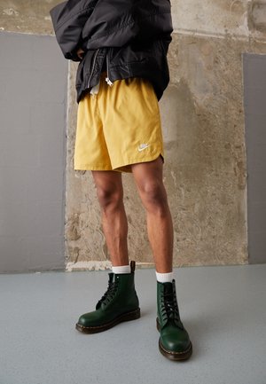 Osoba nosící žluté šortky Nike, tmavě zelené šněrovací boty, bílé ponožky a černou bundu, stojící na šedé podlaze před texturovanou zdí.