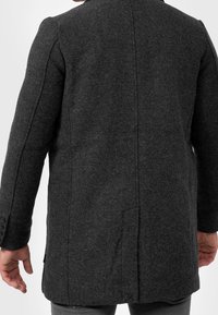 Dunkelgrauer Wollmantel mit Fischgrätenmuster, tailliertem Schnitt, Gehschlitz hinten und dreiknöpfigem Detail an den Ärmeln.