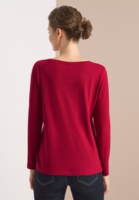 Czerwona, długosleeve T-shirt uszyta z miękkiego materiału, z szerokim dekoltem i luźnym krojem, starannie zestawiona z ciemnymi dżinsami i subtelnymi detalami przeszyć.