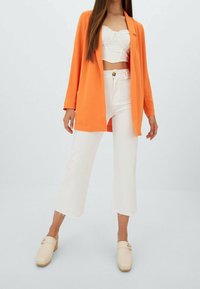 Blazer arancione, top corto bianco e pantaloni bianchi a vita alta. Zoccoli beige con dettagli in metallo dorato. Tessuto morbido in tutto il completo.