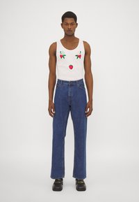 Top blanco sin mangas con fresas y hojas bordadas, combinado con unos vaqueros de mezclilla azul de cintura alta. Unas botas negras de suela gruesa completan el atuendo.