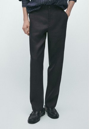 Persona che indossa pantaloni neri a gamba dritta, mocassini neri e una camicia scura a righe sottili con le mani in tasca.