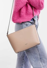 Beige kožená crossbody taška se strukturovaným tvarem, jemným logem, tenkým popruhem a hladkým povrchem. Nosená s světle modrými džíny.