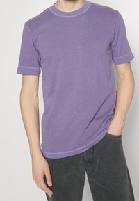 Fioletowa bawełniana koszulka z krótkim rękawem, okrągłym dekoltem i widocznymi detalami szycia. Połączona z ciemnoszarymi dżinsami.