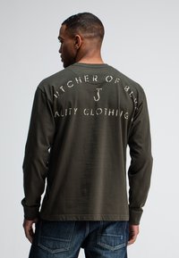 Langärmliges olivgrünes Shirt mit weißem Blocktext "BUTCHER OF BLUES QUALITY CLOTHING" auf dem Rücken. Weicher Stoff, entspannte Passform.