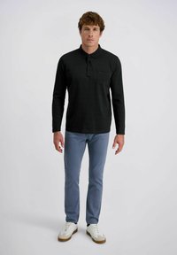 Langärmliges schwarzes Polohemd mit einer subtilen Streifenstruktur, einer einzelnen Brusttasche, kombiniert mit hellblauen Hosen und weißen Sneakers.
