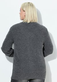 Žena s blond vlasy, která má na sobě stříbrné kruhové náušnice a tmavě šedý ribovaný pletený svetr, zobrazená zezadu na pozadí bílé zdi.
