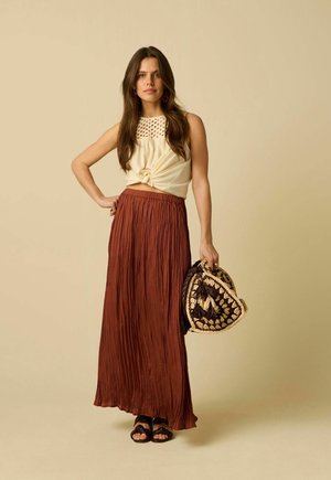 Vrouw met lang bruin haar, gekleed in een mouwloos crèmekleurig gehaakt topje geknoopt in de taille, roestkleurige geplooide maxi rok, sandalen en een geweven tas in de hand.