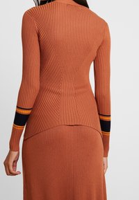 Hladký, těsně přiléhající top s dlouhým rukávem v spálené oranžové barvě, s kontrastními námořnickými modrými a žlutými pruhy na manžetách a výraznými švy na zadní straně.