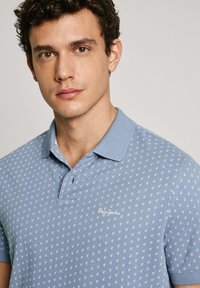 Světle modré polo tričko s vzorovaným designem, na kterém jsou malé bílé kotvy, klasickým límcem a logem na levé straně hrudi.