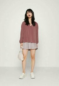 Top largo de mangas compridas em cor bordô com decote em V, combinado com uma saia rodada cinza-claro. A modelo segura uma pequena bolsa branca e usa sapatos de plataforma brancos.