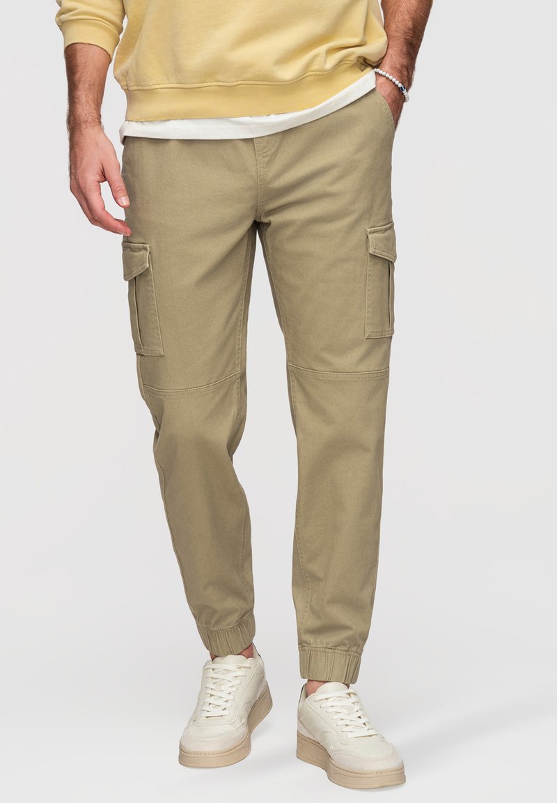 Homme portant un pantalon cargo beige avec des poignets élastiques, des baskets blanches, un sweat jaune et un débardeur blanc, main dans la poche, debout.