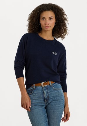 Navy blue knitted sweater features a round neckline and long sleeves, with a subtle embroidered logo on the chest. Worn with blue jeans and a brown belt.