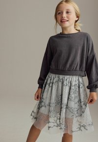 Sweatshirt gris court à manches longues associé à une jupe en tulle grise superposée avec de la broderie argentée et des sequins. S'assoit au-dessus du genou.
