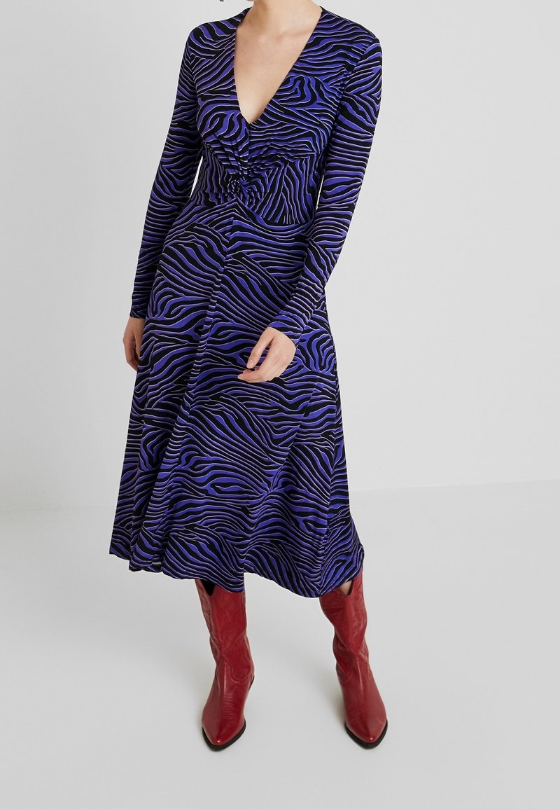 Klänning med långa ärmar och djup V-ringning i blått och svart zebramönster, med draperad detalj vid midjan, kombinerad med röda läderstövlar som går upp till knäna.