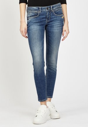 Skinny blue denim jeans with a faded texture, five pockets, and a button closure, paired with white sneakers.