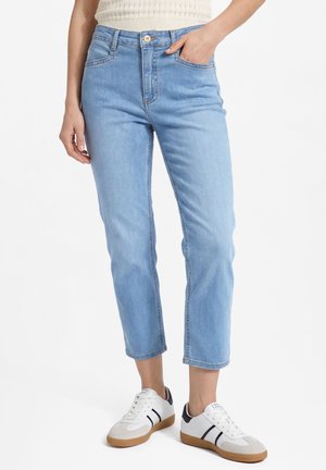 Frau trägt hellblaue kurze Jeans, weiße Sneakers mit schwarzen Streifen und ein cremefarbenes gestricktes ärmelloses Oberteil, steht mit einer Hand in der Tasche.
