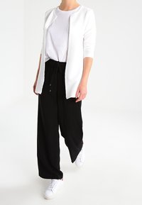 Weiße offene Strickjacke über einem weißen T-Shirt, kombiniert mit lockeren schwarzen Hosen und weißen Turnschuhen. Einfaches Design ohne Muster.