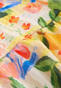 Blomstermønstret stof med gule, lyserøde, blå og grønne nuancer. Tekstureret, letvægtsmateriale med synlige syninger langs sømmene.