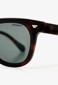Gafas de sol de carey marrón con lentes verdes. Presenta el logo "SUPERDRY" en la parte superior izquierda y un acento dorado en el lateral.
