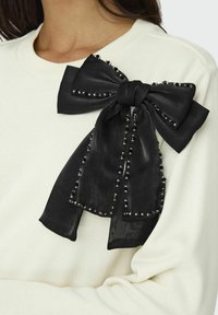 Fiocco di raso nero con accenti di perline attaccato a un maglione color crema. Il fiocco presenta un nodo centrale e estremità fluttuanti, creando un aspetto testurizzato.