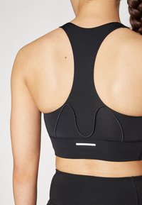 Svart sports-bh med racerback-design, teksturert stoff, dekorativ søm og en liten hvit detalj på baksiden.