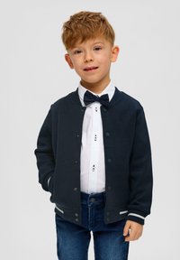 Cardigan blu navy con bottoni a pressione e strisce bianche, indossato sopra una camicia bianca con un papillon e jeans blu. Tessuto liscio, vestibilità casual.