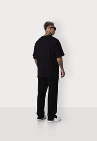 Schwarzes, lässig geschnittenes T-Shirt mit kurzen Ärmeln, kombiniert mit schwarzen Hosen. Das Modell trägt Sonnenbrillen. Der Hintergrund ist einfarbig weiß.