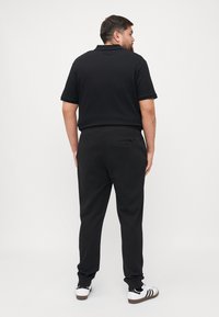 Zwarte polo met een gestructureerd patroon, gecombineerd met zwarte broeken en witte sneakers met zwarte accenten. Weergave van achteren.