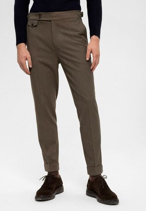 Pantalon marron sur mesure en tissu texturé, avec un devant plat, une fermeture à deux boutons et des revers roulés. Associé à des chaussures foncées.