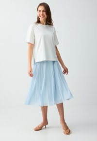 Femme souriante portant un haut blanc à manches courtes, une jupe midi plissée bleu clair et des sandales à talons beiges, debout sur un sol blanc.