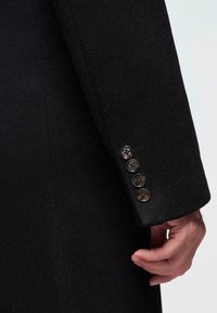 La manga del abrigo de lana negra presenta una textura suave con cuatro botones decorativos plateados, cada uno con un diseño intrincado. Mano descansando suavemente sobre la tela.