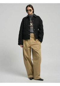 Giacca nera trapuntata con bottoni a pressione, abbinata a un maglione grigio con motivo e pantaloni beige a vita alta. Occhiali da sole sottili completano il look.