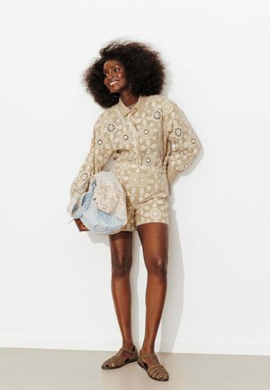 Femme souriante avec une coiffure afro naturelle portant une combinaison beige à manches longues à motifs floraux, tenant un sac en dentelle bleu et blanc, debout.