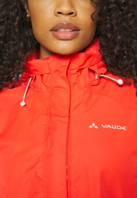 Red waterproof jacket with a high collar, adjustable drawstrings, and a logo. Smooth texture and breathable fabric visible.