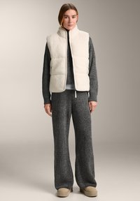 Cremefarbene, flauschige Weste über einem dunklen Strickpullover, kombiniert mit weitem grauen Hose und beigen Schlupfschuhen. Schlichtes Design, keine Muster.