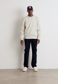 Helles beige Sweatshirt, marineblaue Hose und weiße Sneakers. Die Kappe hat eine blaue Basis mit roten und weißen Akzenten. Schlichtes Design, lockere Passform.