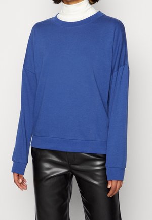 Person iført en blå langærmet sweater over en hvid rullekrave og sorte læderbukser, mod en ensfarvet baggrund.