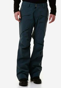 Pantaloni impermeabili di un verde scuro con vestibilità ampia, molteplici tasche e cerniere alle caviglie. Presentano una chiusura a bottone e un tessuto testurizzato.