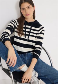 Jeune femme aux longs cheveux bruns assise sur une chaise en métal, portant un sweat à capuche rayé noir et blanc, un jean bleu, des chaussettes blanches et des baskets grises.