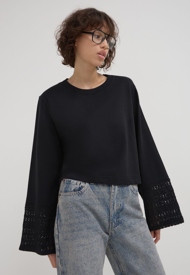 Felpa nera corta con maniche lunghe e svasate, caratterizzata da dettagli in pizzo elaborati nei polsini, abbinata a jeans in denim blu chiaro.