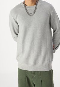 Felpa grigia con texture, dal collo a giro e orlo a costine. Abbinata a pantaloni verdi, con due catene sovrapposte come accessori.