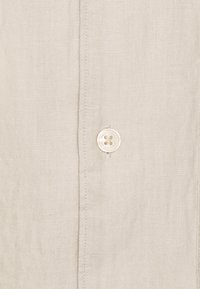 Lichtbeige linnen overhemd close-up, met een ronde, crèmekleurige knoop met vijf gaatjes, verticale stiksels en een gestructureerde geweven stof.